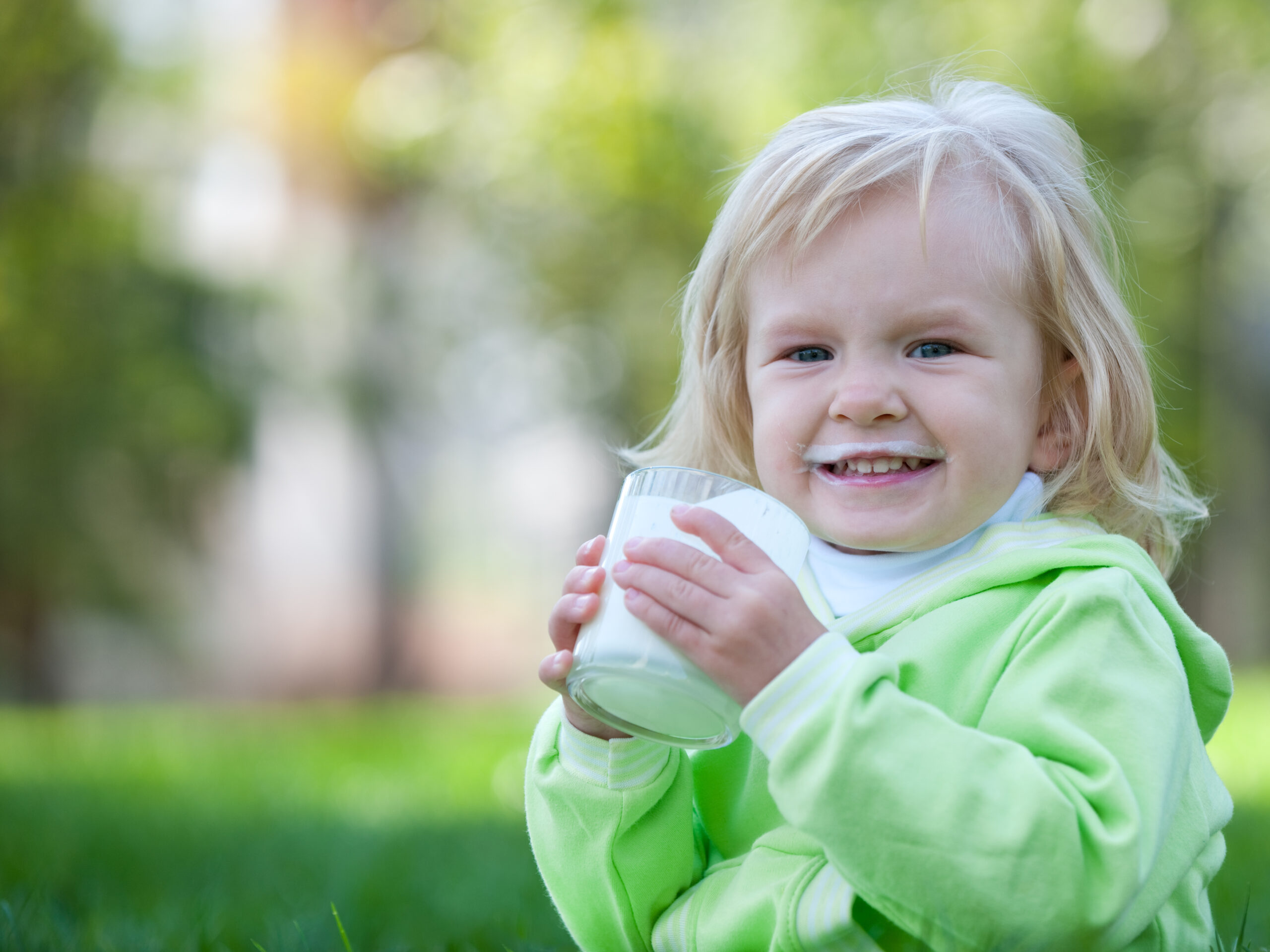 Blonde toddler drinking a glass of milk in the garden outdoors