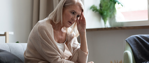 An older woman sitting on her lounge with a concerned look on her face holds her hand to her head.